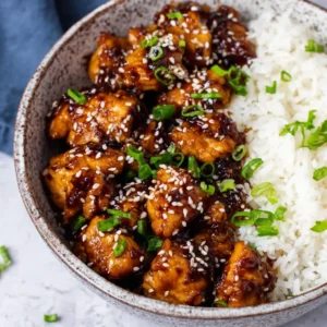 Easy healthy sesame chicken is served in a white bowl with rice and garnished with sesame seeds and green onions, showcasing a delicious and simple weeknight meal.
